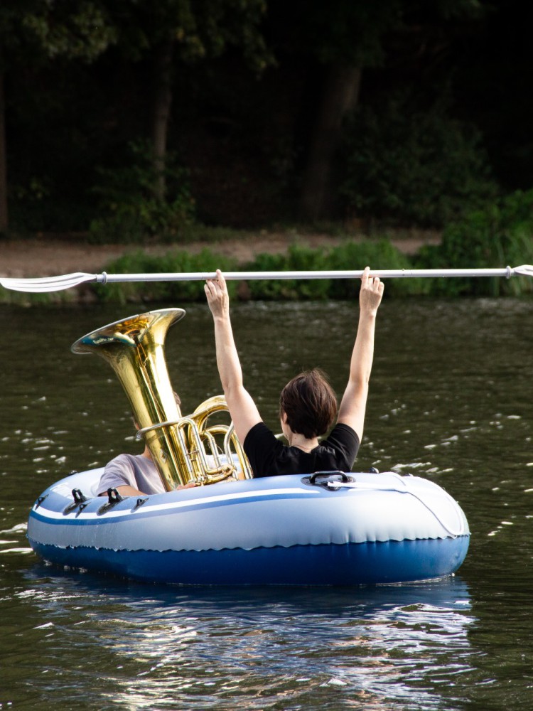 Ein Schlauchboot im Wasser, darin eine Person versteckt hinter einer Tuba, eine weitere hat ein Paddel in ihren nach oben gestreckten Armen