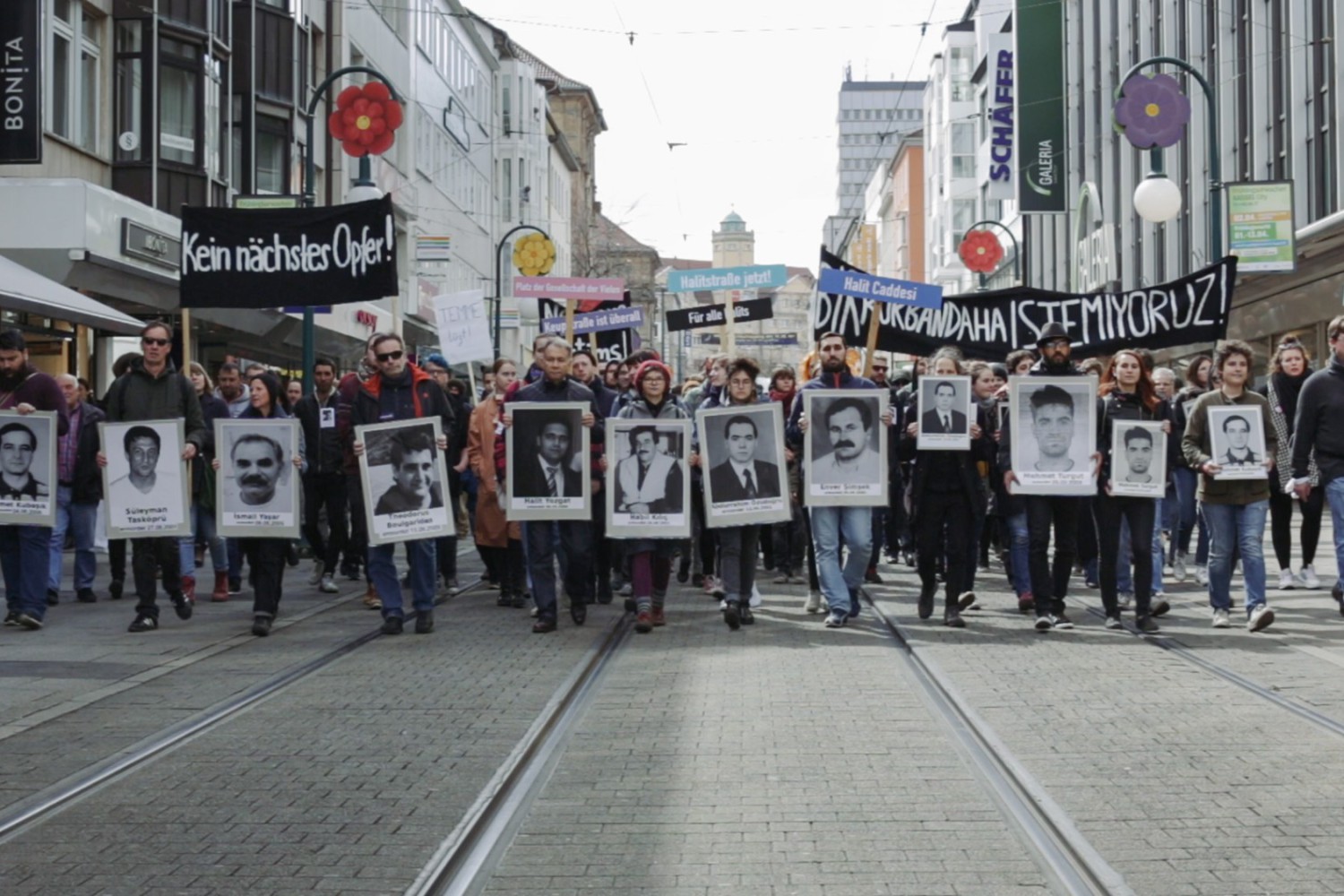 Szene aus dem Film "Der zweite Anschlag", in der man eine Gruppe von Menschen sieht mit Schildern, die an Ermordete erinnern. Die Menschen laufen in einer Demonstration auf uns zu.