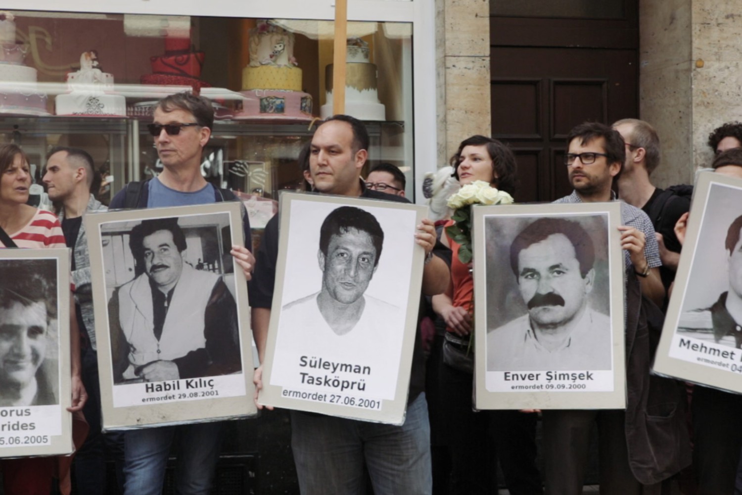 Szene aus dem Film "Der zweite Anschlag", in der man eine Gruppe von Menschen sieht mit Schildern, die an Ermordete erinnern.