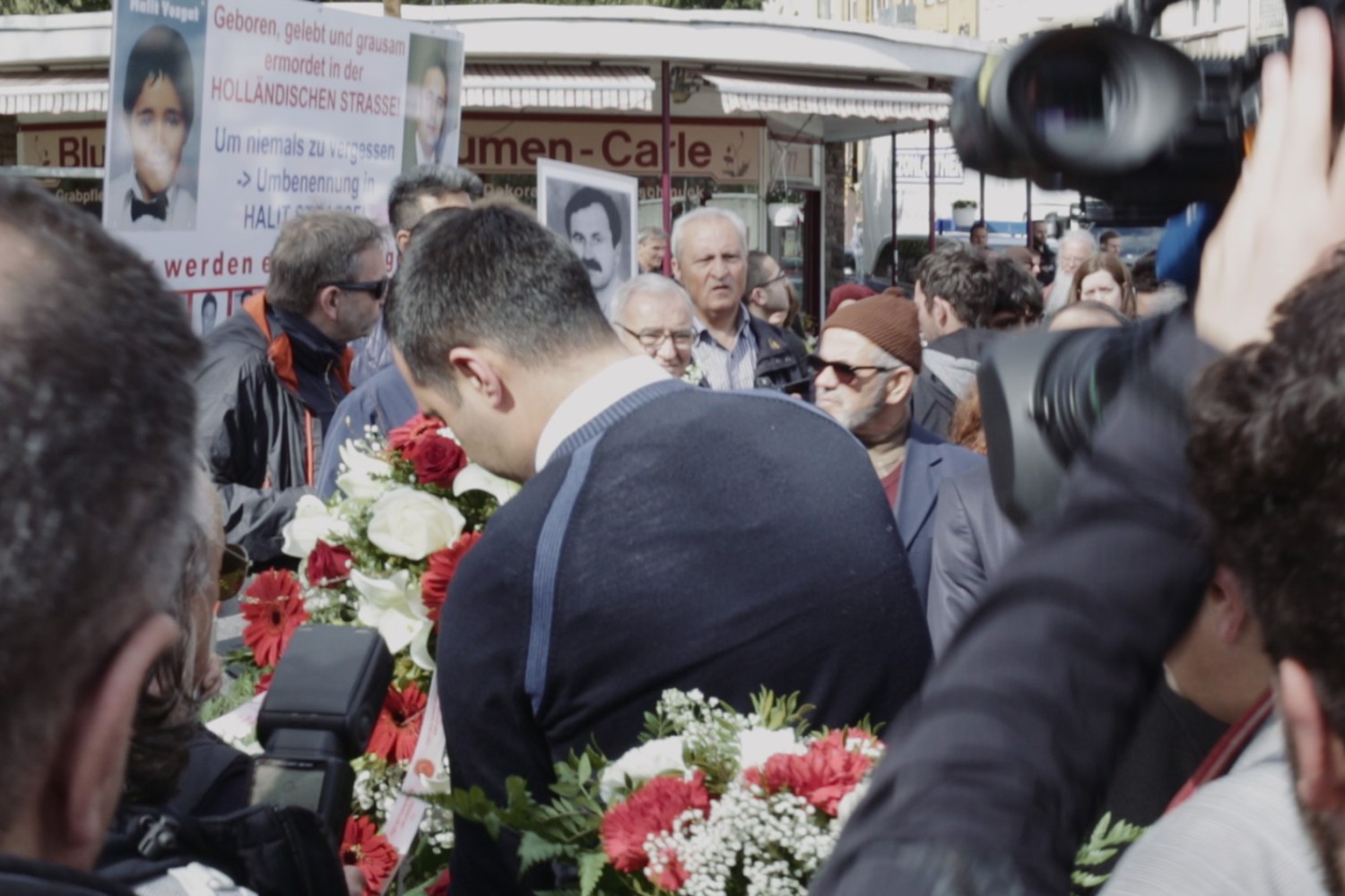 Szene aus dem Film "Der zweite Anschlag", in der man eine Gruppe von Menschen sieht mit Schildern, die an Ermordete erinnern.