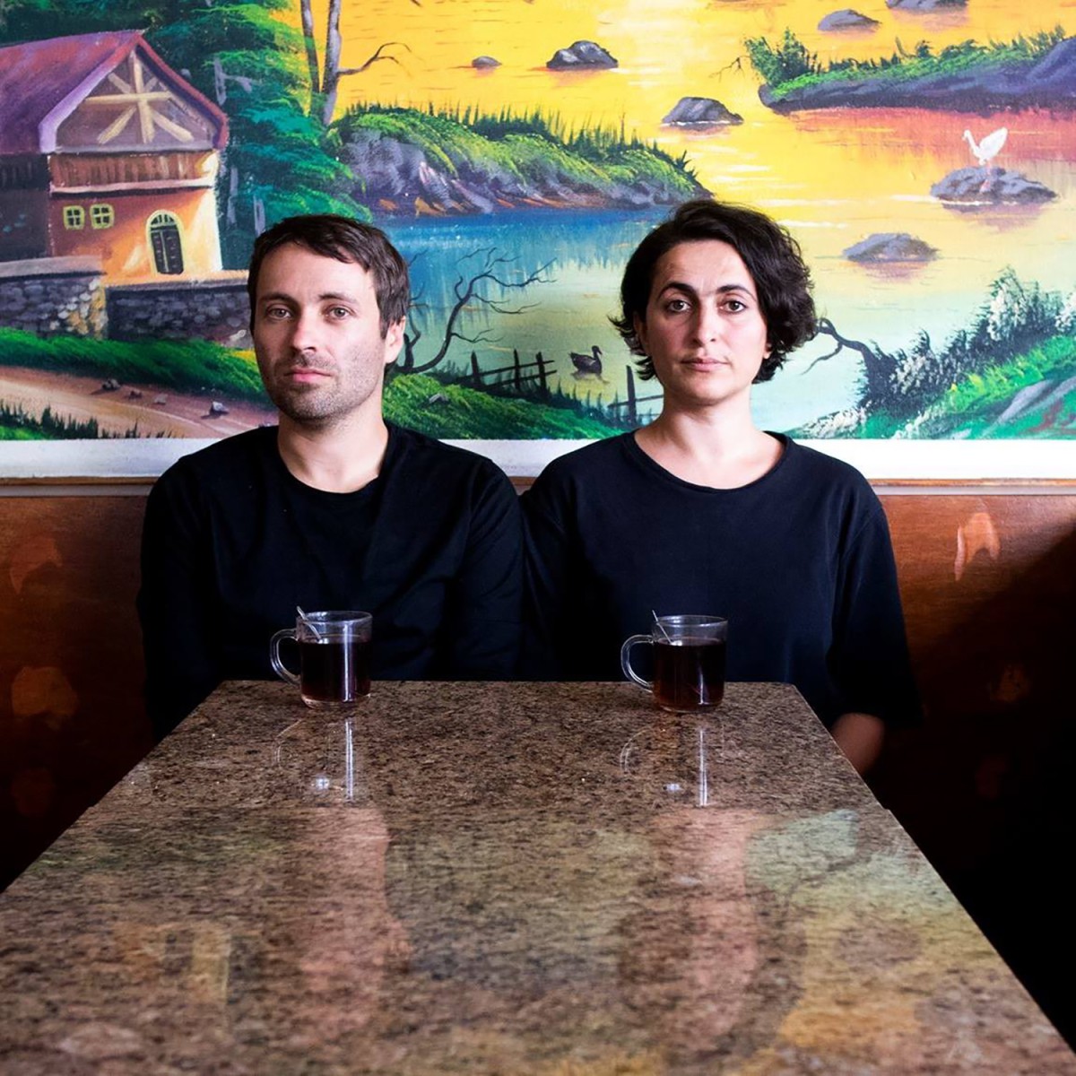 Nuray Demir and Michael Annoff sit next to each other at a table, tea in front of them, a fairy tale wallpaper behind them