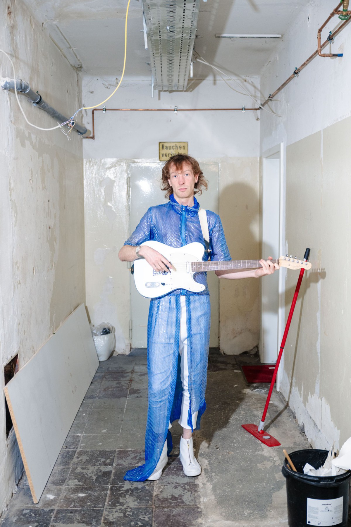 TCHAIR mit weißer Gitarre und blauem Pailetteneinteiler in einer Abstellkammer