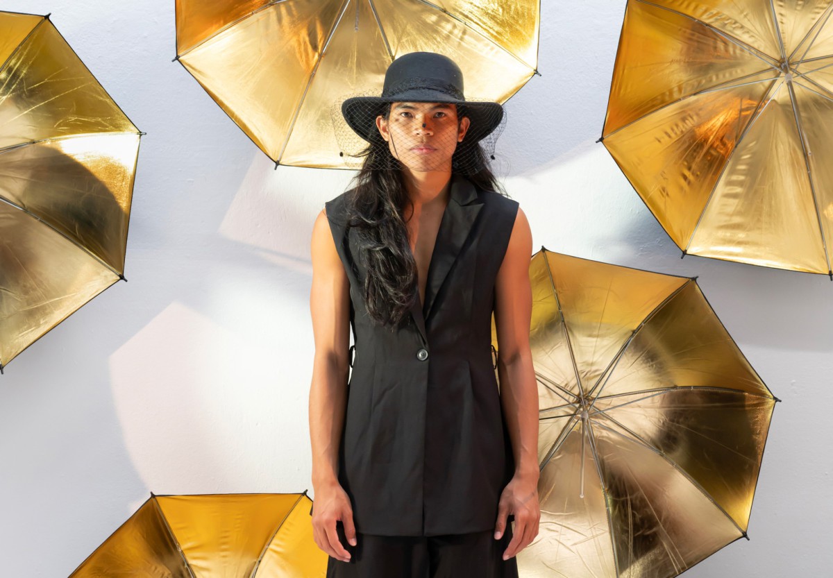Der Tänzer Jed Nagales in einem schwarzen ärmellosen Anzug, im Hintergrund aufgespannte goldene Regenschirme